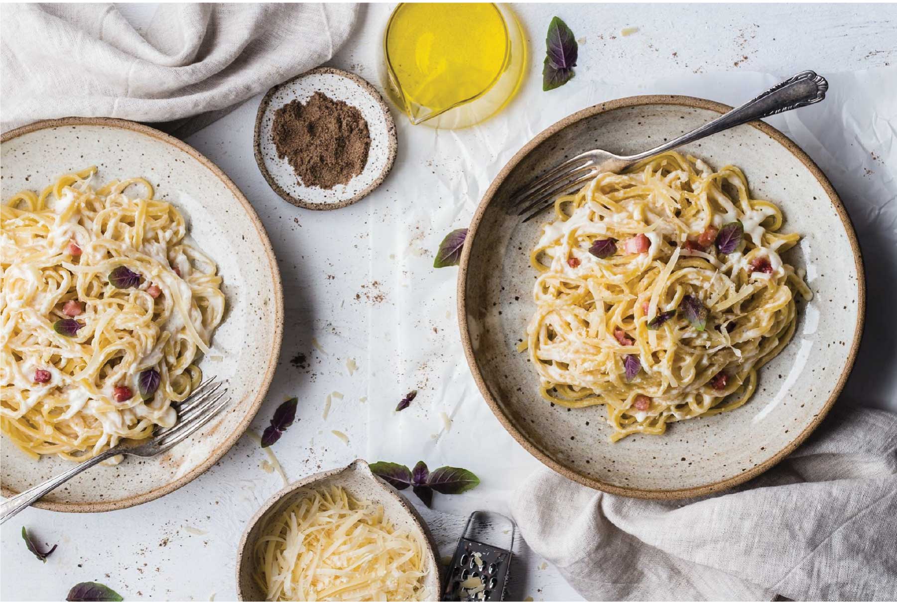 two beige plates of pasta carbonara on a white cloth background