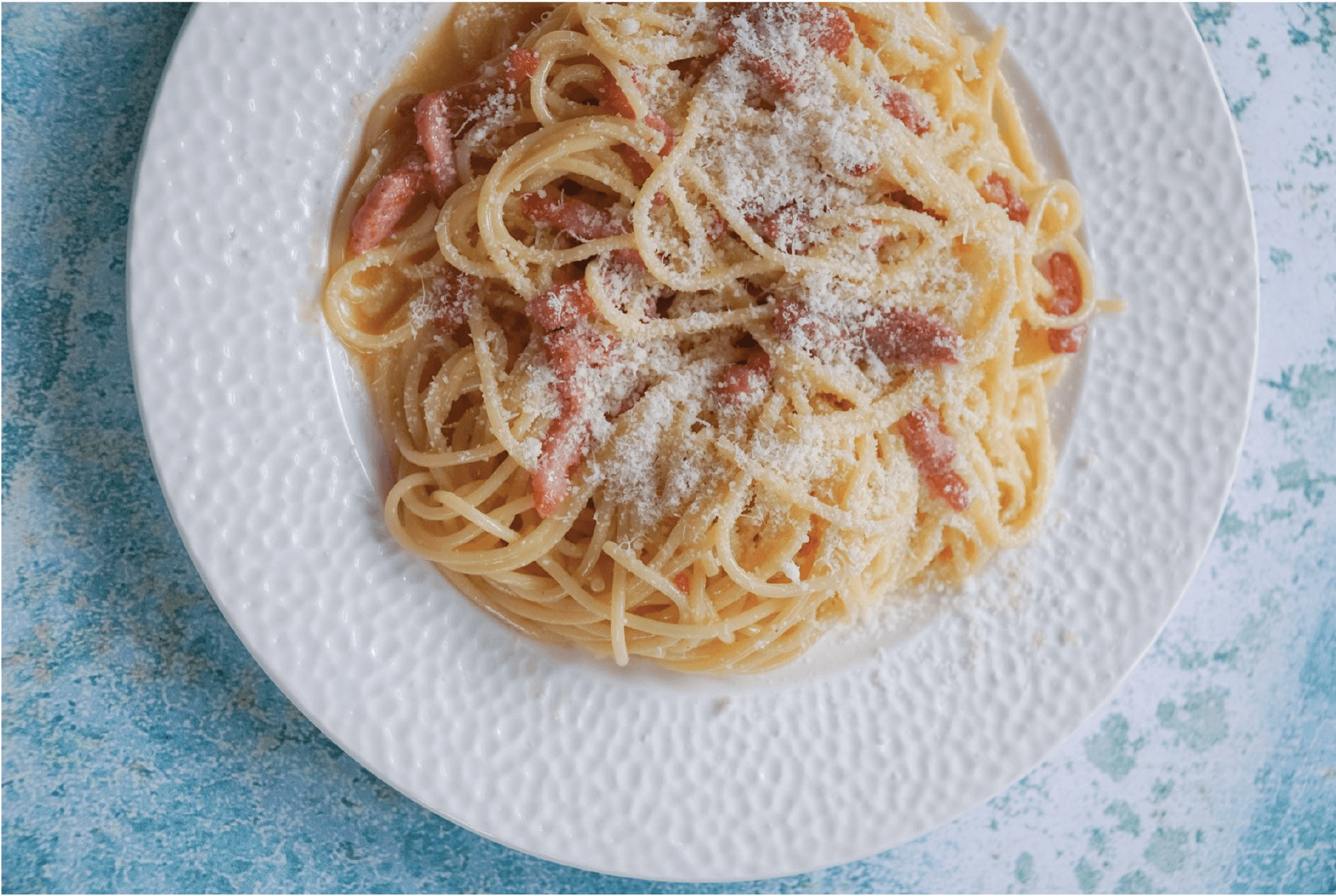 pasta carbonara topped with bacon and parmesan on a white plate, with a blue background